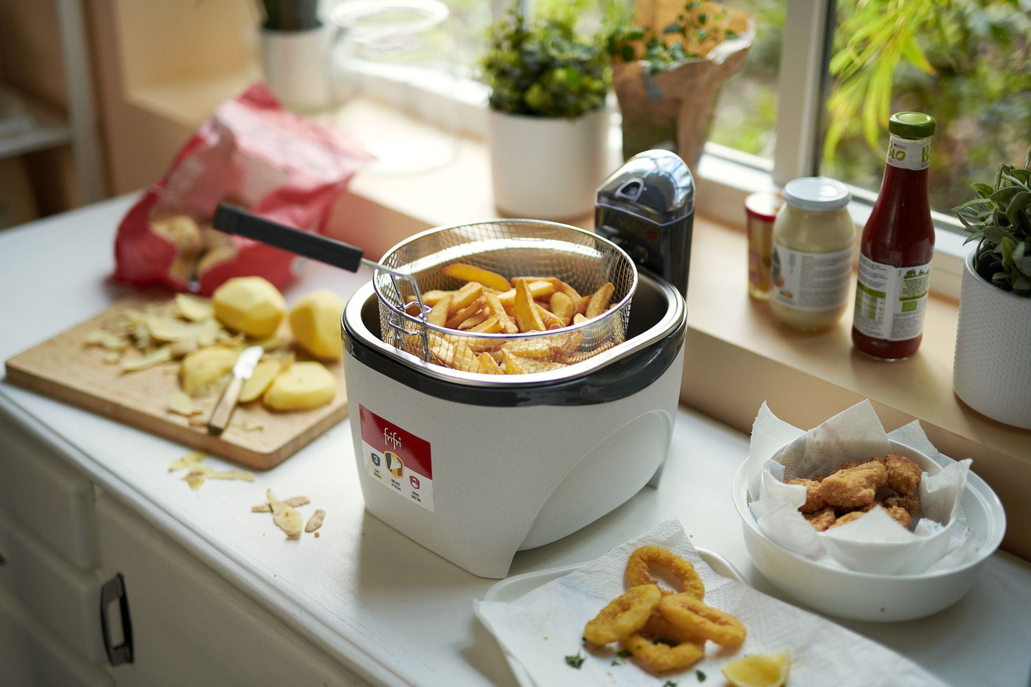 Une friteuse avec des frites dans le panier est posée sur le comptoir d'une cuisine à côté d'un bol d'aliments frits, de condiments et d'une planche à découper avec des tranches de pommes de terre.