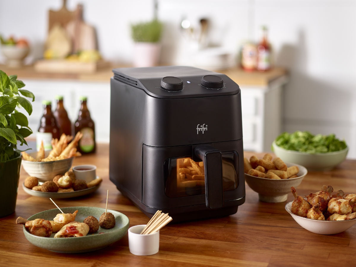 Une friteuse noire est posée sur un comptoir de cuisine, entourée d'assiettes de snacks frits, de sauces à tremper et de bouteilles, avec des ustensiles de cuisine en arrière-plan.