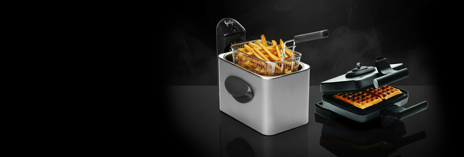 Een zilveren friteuse met een mand frietjes naast een zwart wafelijzer met twee gebakken wafels, beide met zichtbare stoom, op een zwart glanzend oppervlak.