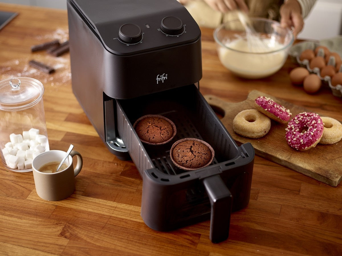 Un panier de friteuse contenant deux muffins au chocolat est posé sur un comptoir en bois, avec en arrière-plan des beignets, des œufs, du sucre et une personne en train de mélanger de la pâte.