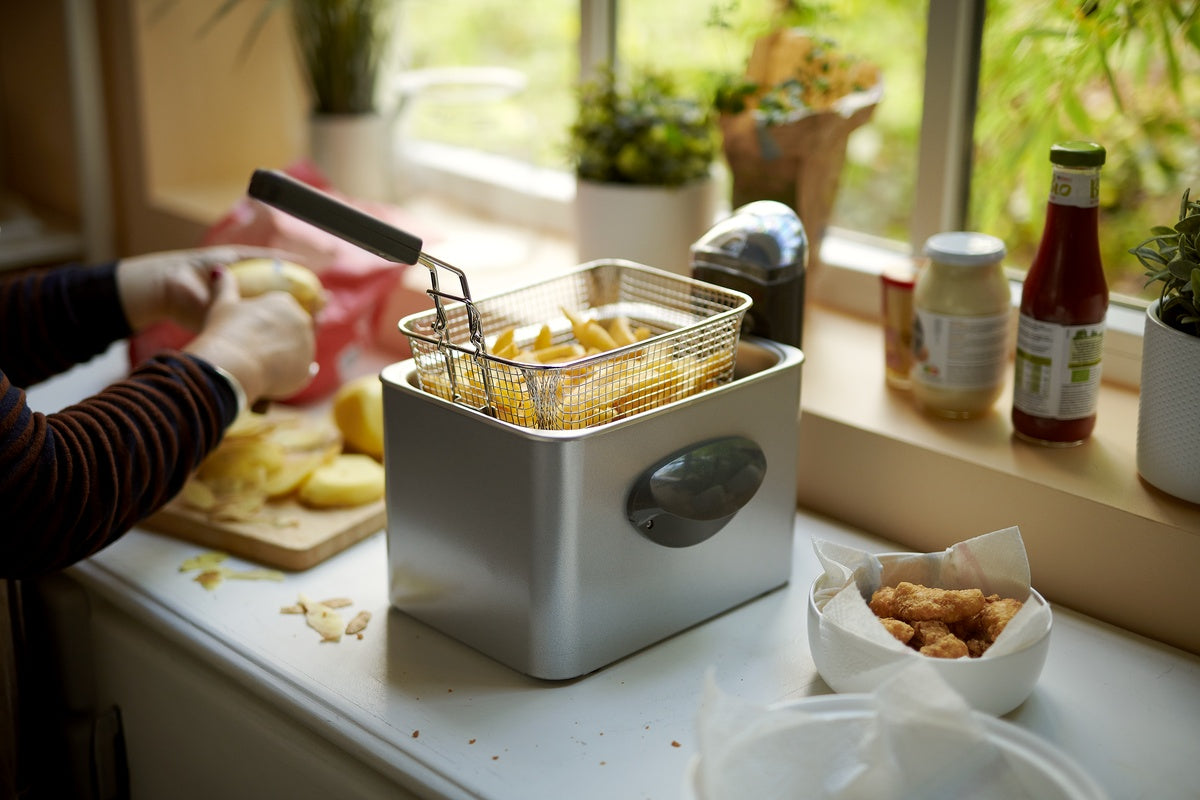 Een persoon schilt aardappelen naast een friteuse met aardappelschijfjes erin, terwijl een kom gefrituurde voedingswaren in de buurt op een keukenblad staat.