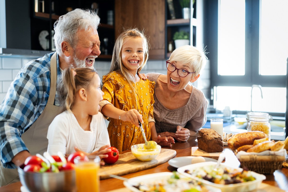 Twee volwassenen en twee kinderen staan samen in een keuken, glimlachend en etend aan het bereiden met diverse schalen en ingrediënten op het aanrecht.