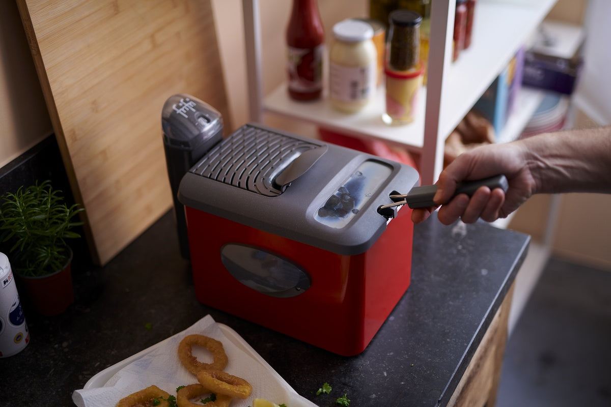 Une personne utilise des pinces pour retirer la glace d'une machine à glaçons de comptoir rouge, avec des rondelles d'oignon et des condiments visibles à proximité.