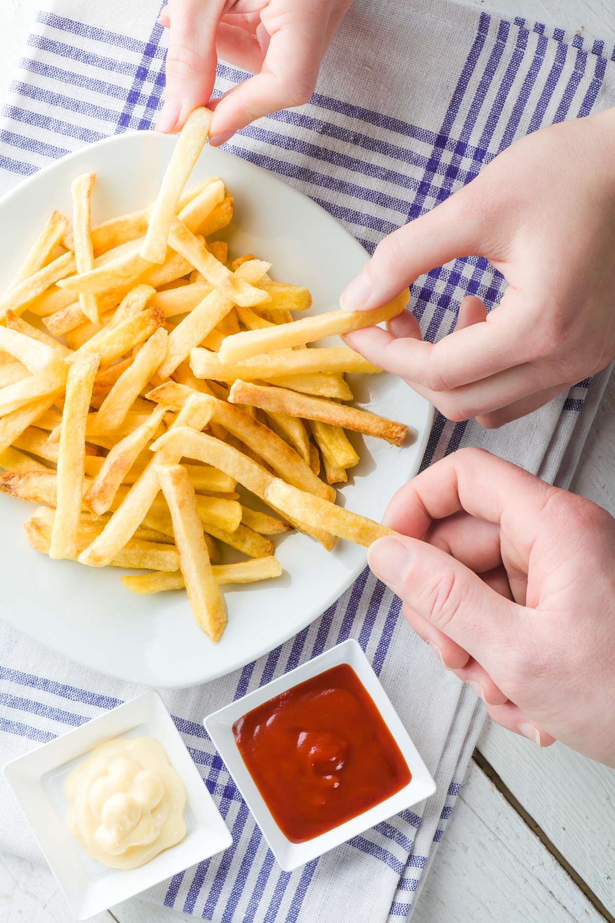 Deux personnes prennent des frites sur une assiette, avec des petites coupelles de ketchup et de mayonnaise sur le côté, posées sur une serviette en tissu rayé.