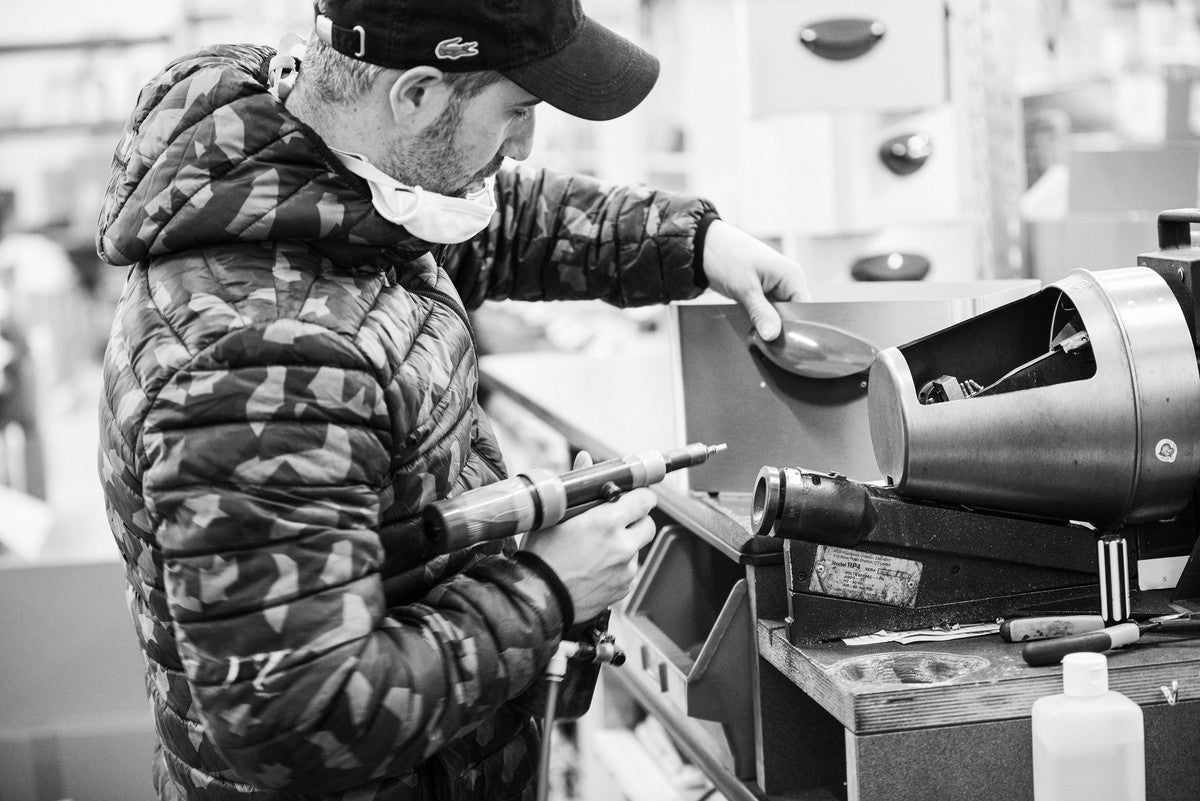 Un homme portant une casquette et une veste utilise un outil électrique pour travailler sur une machine à un établi. Des outils et des équipements sont visibles autour de lui.