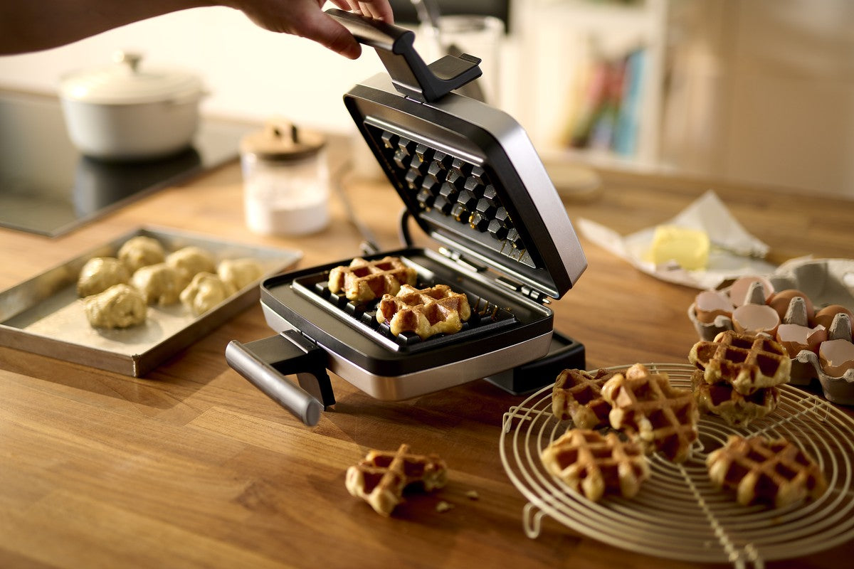 Een persoon tilt het deksel op van een wafelijzer met verse wafels erin; deegballetjes, eieren en gebakken wafels liggen op het houten aanrecht in de keuken.