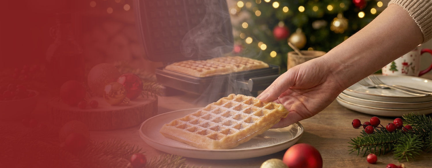 Een hand legt een met poedersuiker bestrooid wafel op een bord naast een wafelijzer, met kerstdecoraties en een kerstboom op de achtergrond.