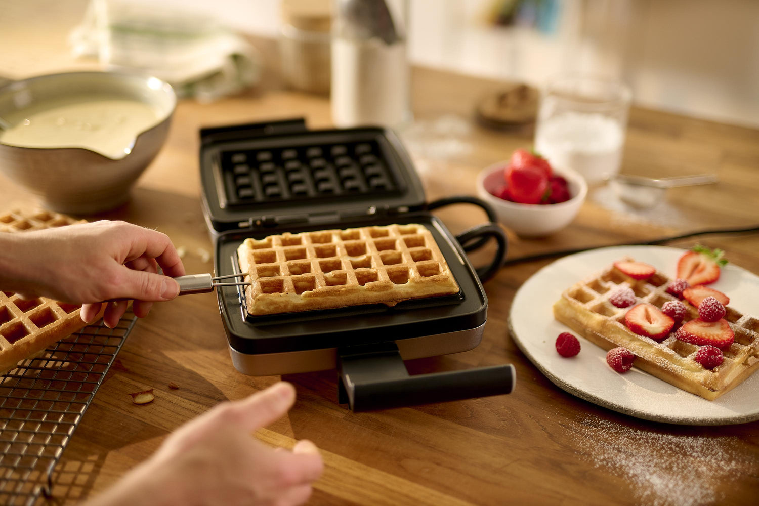 Une personne retire une gaufre fraîchement cuite d'un gaufrier ; une assiette contenant des gaufres et des fraises est posée à proximité sur un comptoir en bois.