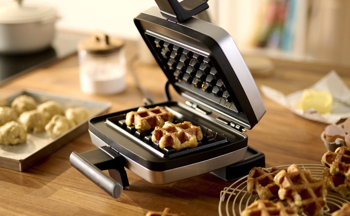 Een open wafelijzer met gedeeltelijk gebakken wafels ligt op een houten aanrecht, omgeven door deegballetjes, een afkoelrek en keukengereedschap.