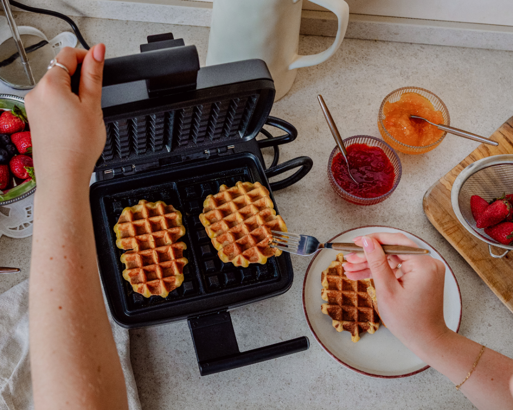 Une personne utilise une fourchette pour retirer une gaufre cuite d'un gaufrier ; à côté, sur le plan de travail, se trouvent des bols de confiture et de fraises fraîches.