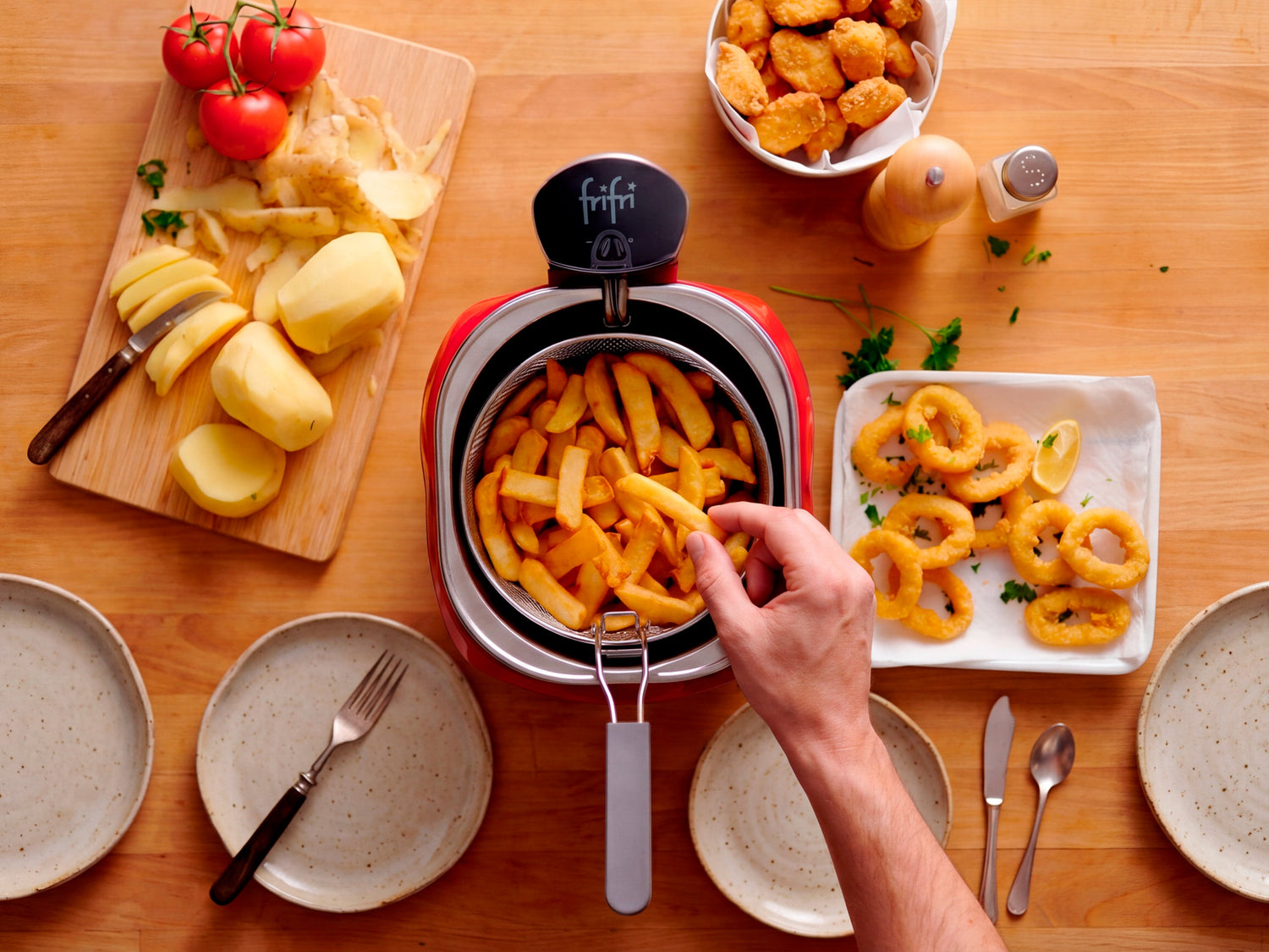 Une main sort des frites tout juste cuites d'une friteuse posée sur une table où sont disposés des assiettes vides, des légumes coupés, des nuggets dorés et des calamars frits.