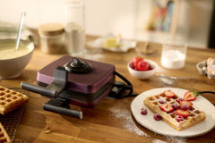 Un Gaufrier Violet + 8 plaques est posé sur une table en bois à côté d'une assiette de gaufres à la fraise et à la framboise ; des ingrédients de cuisson assortis apparaissent à l'arrière-plan.