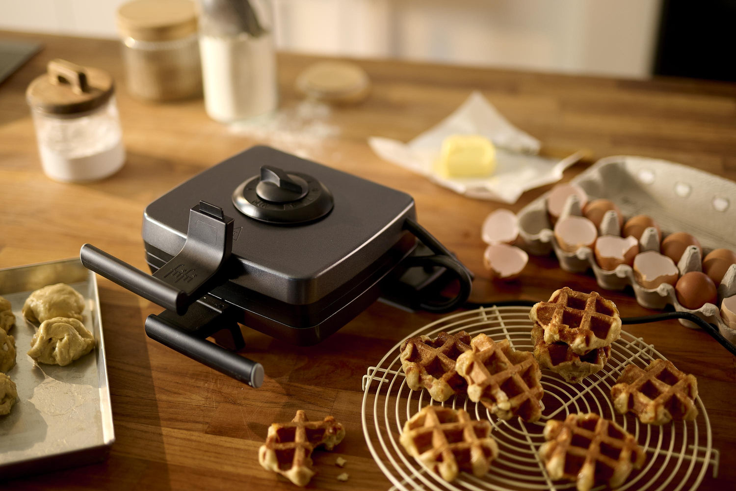 Een Zwart Wafelijzer + 8 platen staat op een houten aanrecht met eieren, boter, bloem, beslag en warme wafels op een afkoelrek – een perfecte aanvulling op elke thuiskeuken.