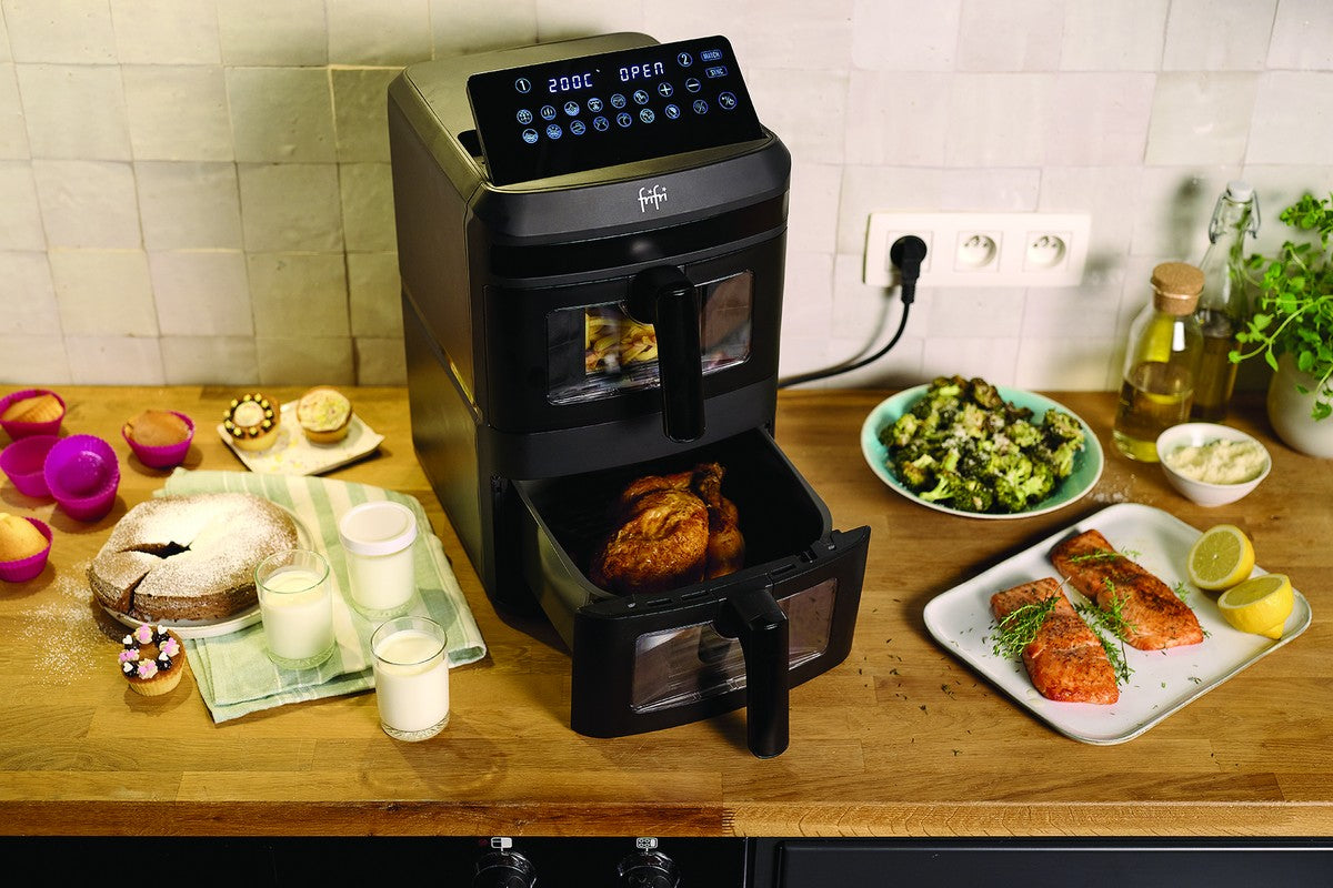 Une friteuse à deux tiroirs est posée sur un comptoir de cuisine avec du poulet cuit à l'intérieur ; des assiettes de saumon, des brocolis, des desserts et des boissons sont disposés à proximité.