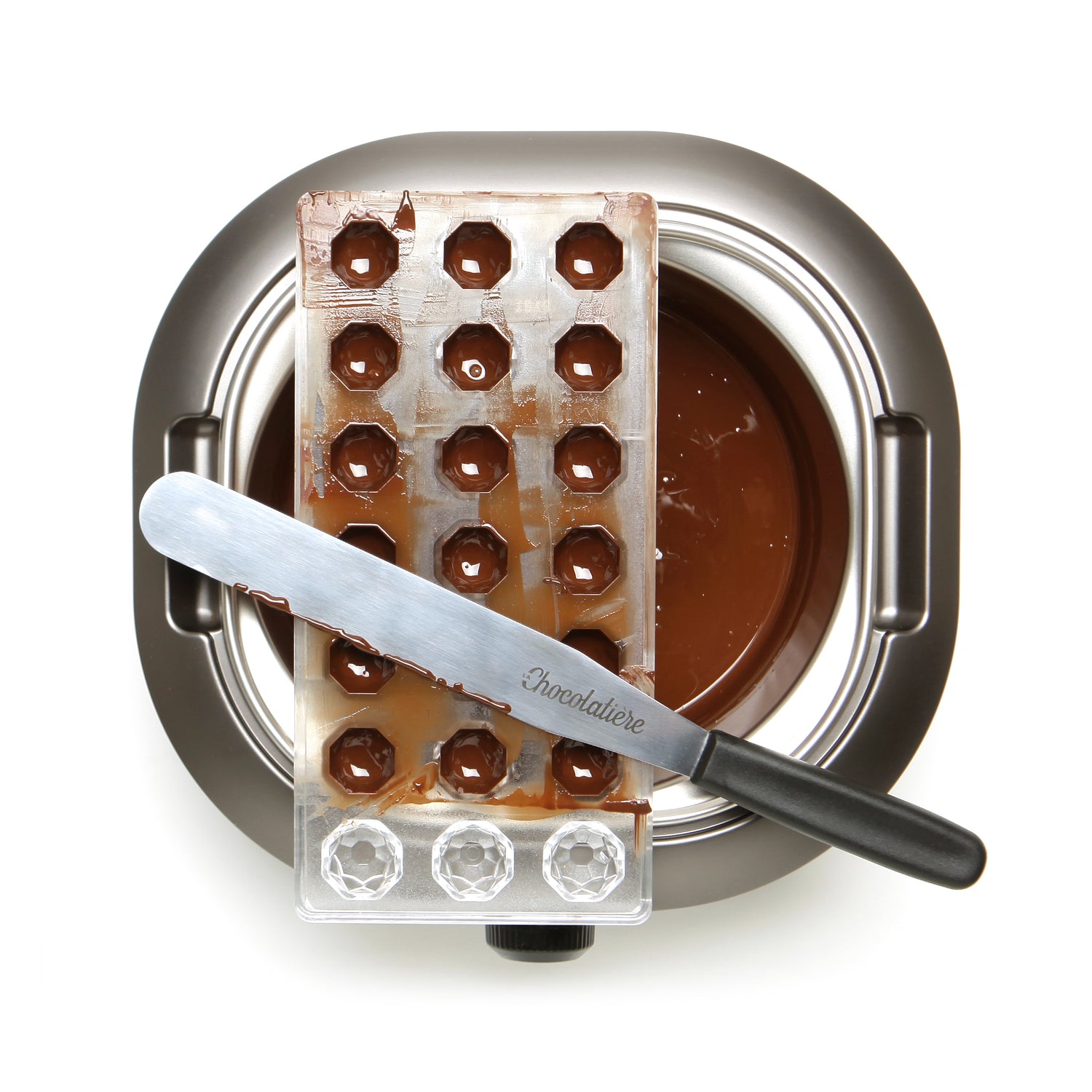Een giettray van De Chocolatière en een spatel liggen op een kom gesmolten chocolade, gezien van bovenaf.