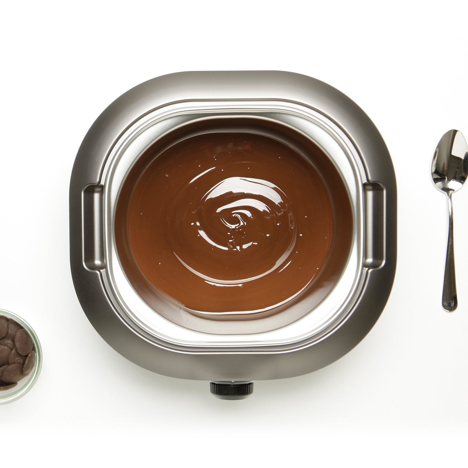 Bovenaanzicht van De Chocolatière + Paaskit gevuld met gesmolten chocolade, naast een lepel en een klein kommetje chocoladestukjes op een wit oppervlak.
