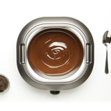 Vue aérienne du chocolat fondu dans un fondoir électrique La Chocolatière en acier inoxydable, avec une cuillère et un petit bol de pépites de chocolat à proximité.