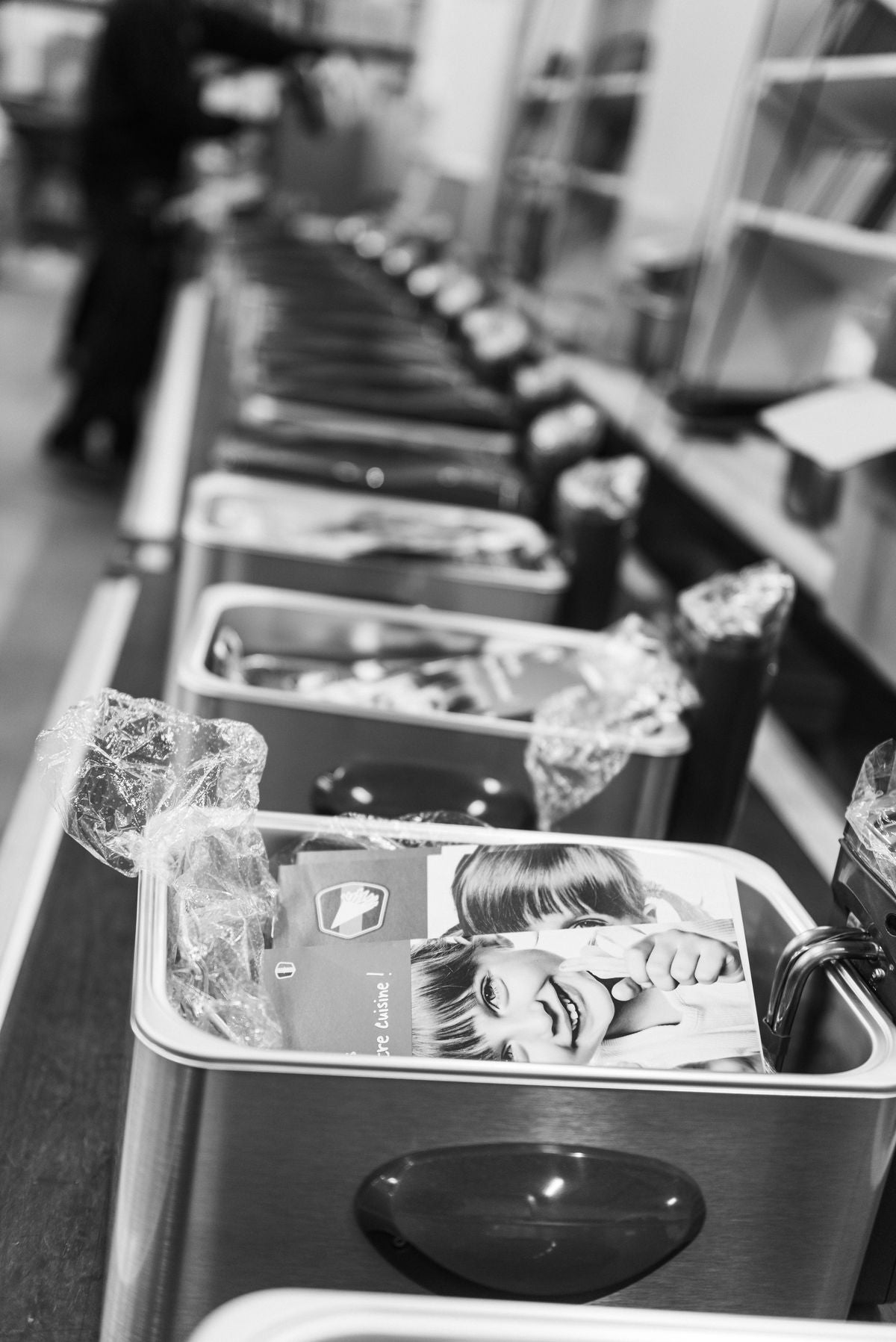 Een rij metalen voedselcontainers met plastic deksels, elk voorzien van een etiket met een lachend kind, geplaatst op een aanrecht in een eenkleurig decor.