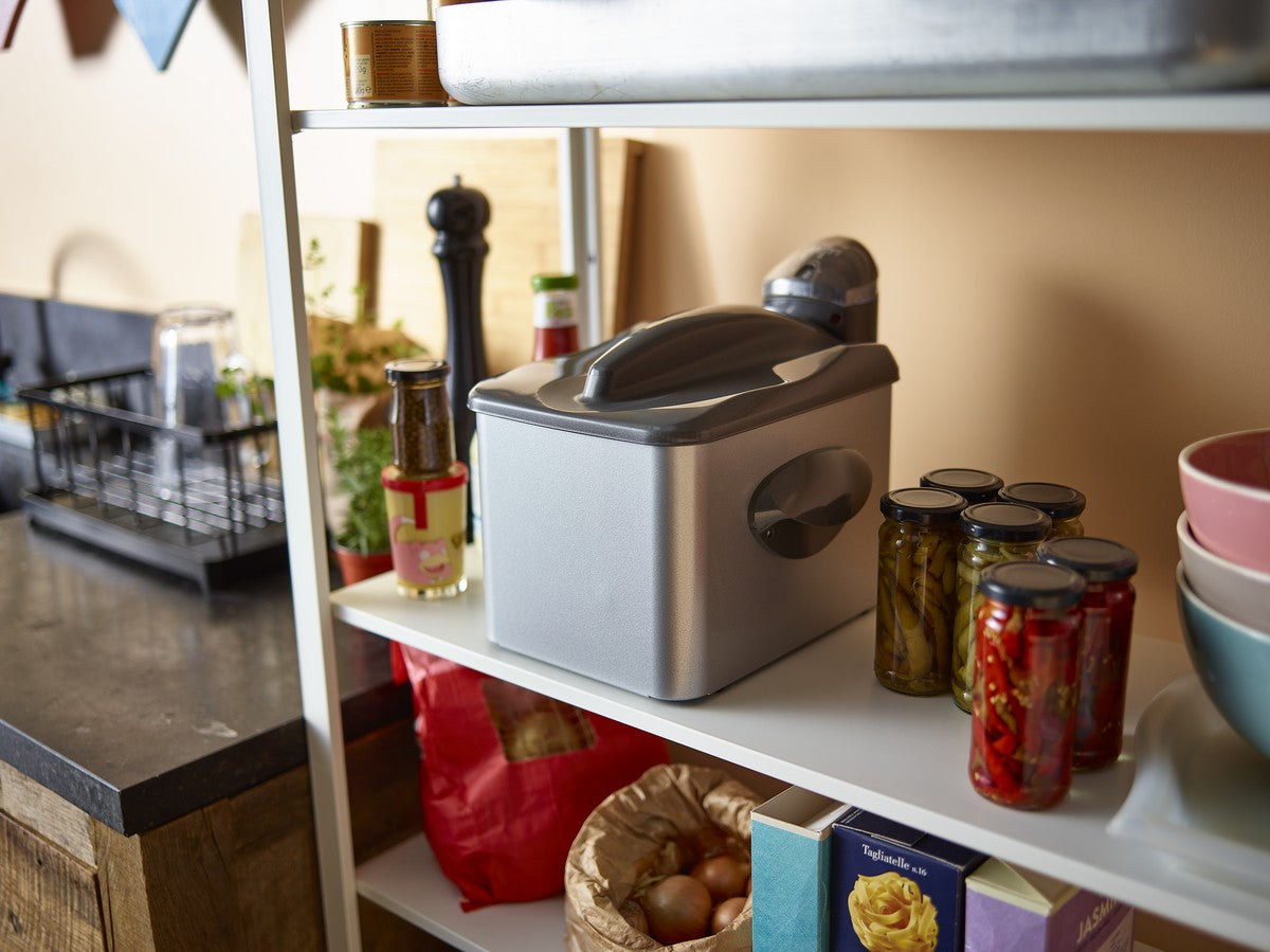 Une huche à pain grise est placée sur une étagère de cuisine blanche, entourée de bocaux de conserves, de bols et de divers ustensiles de cuisine.