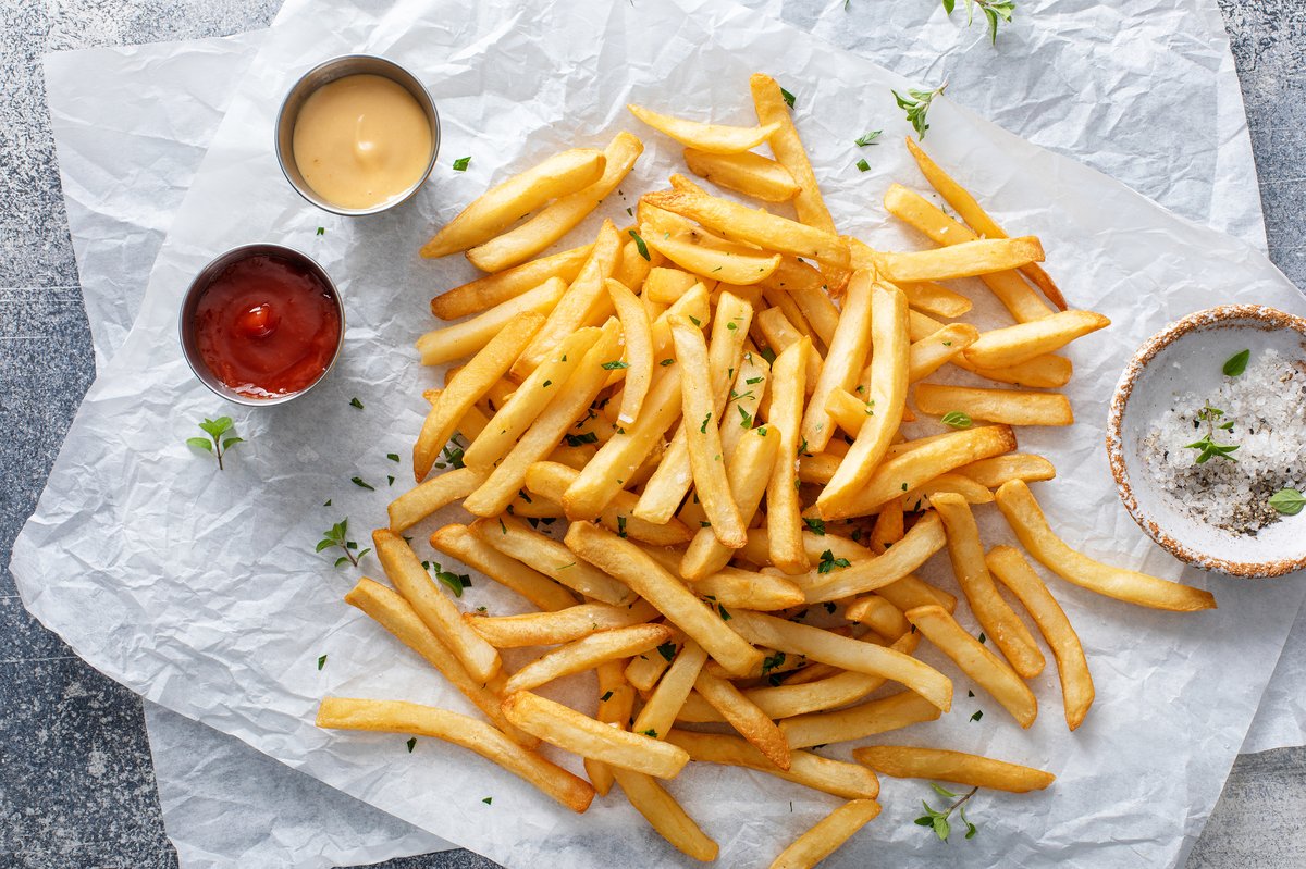 Une portion de frites sur du papier sulfurisé avec un bol de sel, de ketchup et de mayonnaise à côté.