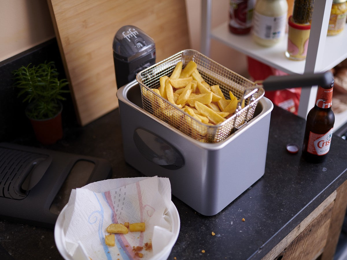 Une friteuse contenant des frites cuites dans un panier métallique est posée sur le comptoir d'une cuisine, à côté d'une assiette contenant quelques frites et un essuie-tout. Une bouteille de bière et des condiments sont également visibles.
