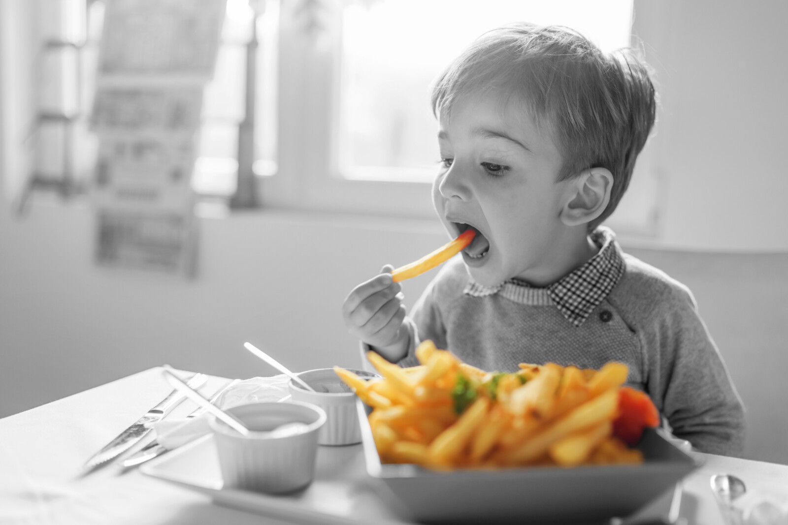 Portrait of small little cute caucasian boy kid eating french fries potato chips at the table in the restaurant or at home three or four years old